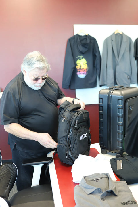 Woz examining a black WOZ-branded OGIO backpack beside luggage.