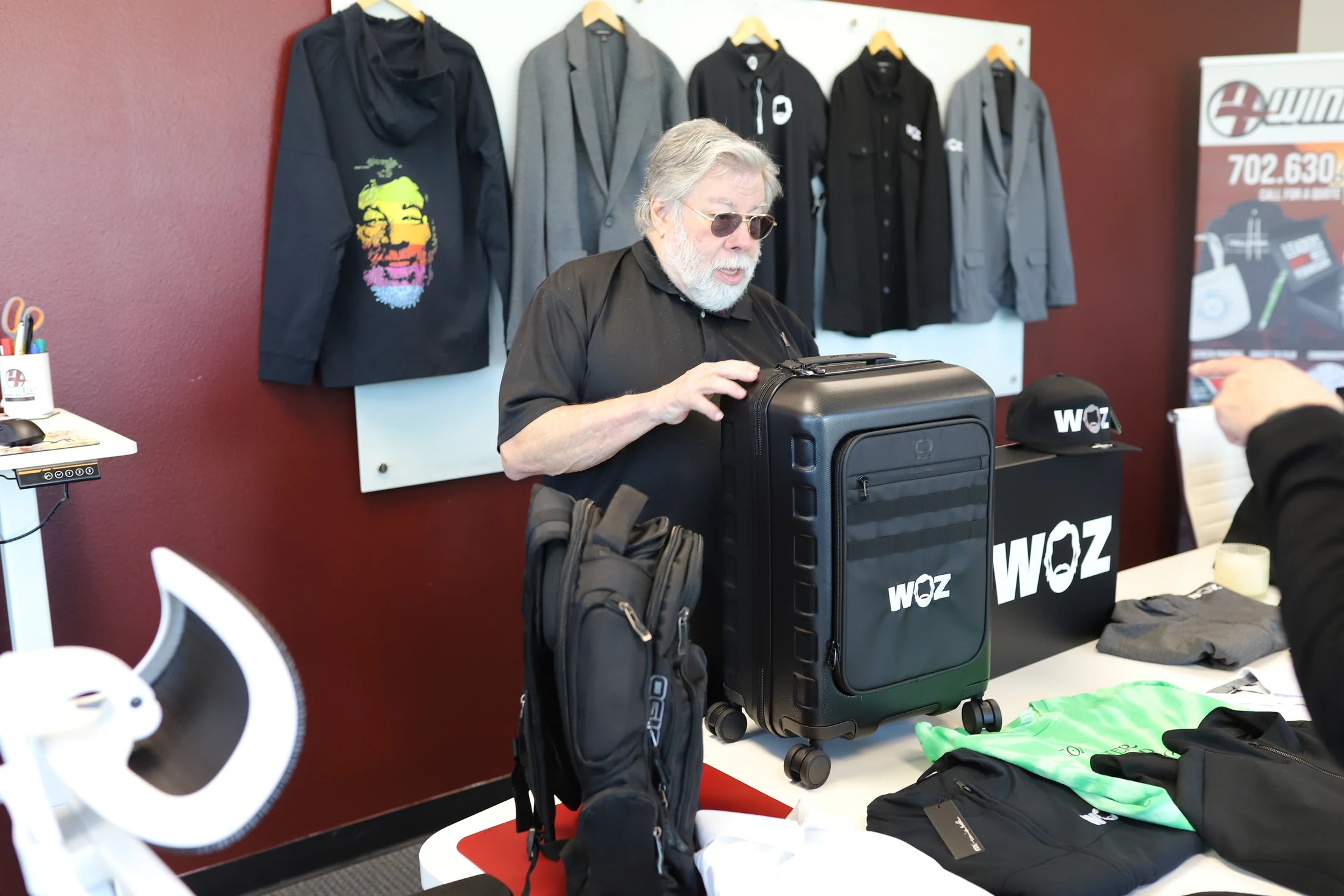 Woz examining black WOZ luggage with apparel displayed behind him.