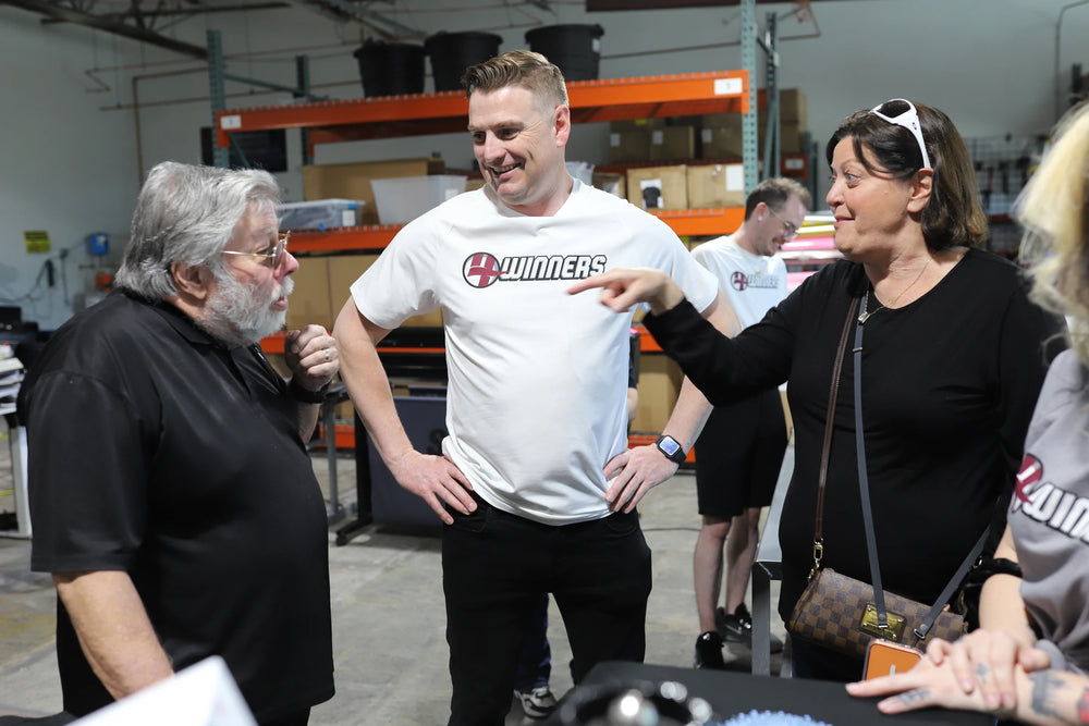 Woz talking with visitors beside production-area shelving.