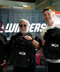 Woz and visitors holding black WOZ-branded OGIO backpacks at the 4Winners facility.
