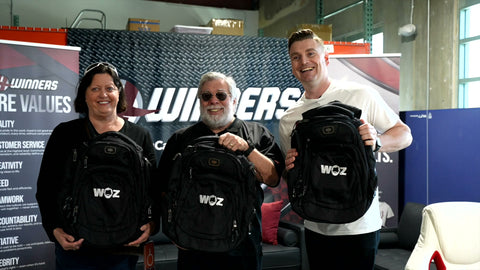 Woz and visitors holding black WOZ-branded OGIO backpacks at the 4Winners facility.