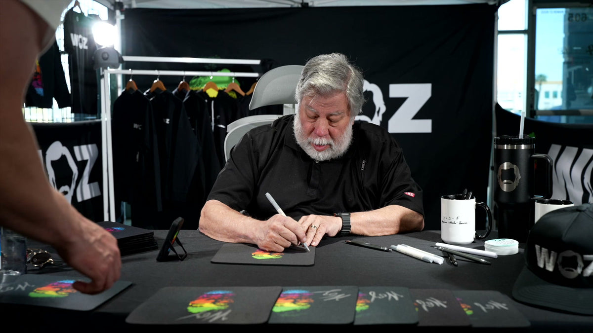 Steve Wozniak signing a black WOZ Rainbow Mouse Pad at the merchandise table during his facility visit.
